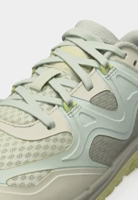 Κοντινό πλάνο από κορδόνια αθλητικού παπουτσιού σε ανοιχτό πράσινο και μπεζ χρώμα πάνω σε αεριζόμενο πλεκτό ύφασμα με συνθετικές επικαλύψεις και υφή στη σόλα.