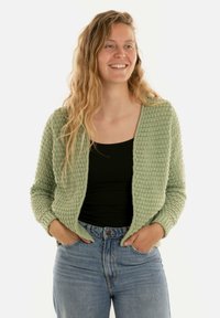 Lichtgroene gebreide cardigan met een gestructureerd patroon, open voorkant, lange mouwen en een relaxte fit, gecombineerd met een zwarte top en high-waisted jeans.