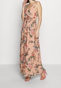 Donna in un maxi dress senza maniche di colore pesca con motivo floreale e scollatura a V, che indossa sandali con tacco alto e fasce corallo, in piedi di fronte a uno sfondo bianco.