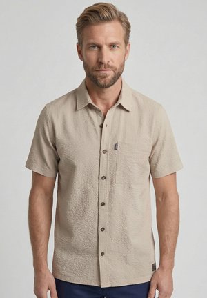 Baardige man met lichtbruin haar die een beige gestructureerd overhemd met korte mouwen en een borstzak draagt tegen een effen achtergrond.