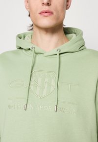 Sudadera verde claro con un logo de GANT texturizado, cordones con terminaciones plateadas y un tejido suave y acogedor. Cuenta con un bolsillo tipo canguro.