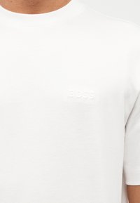 Κοντινό ενός ατόμου που φοράει απλό λευκό T-shirt με μικρό ανάγλυφο λογότυπο «BOSS» στο αριστερό στήθος.