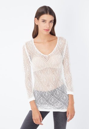 Mujer con el cabello castaño liso, vistiendo una blusa tejida blanca de manga larga y pantalones oscuros, de pie frente a un fondo blanco liso.