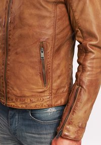 Giacca in pelle marrone con design aderente, cuciture visibili, due tasche con zip e una texture spazzolata. Accentuata da dettagli in metallo argentato.