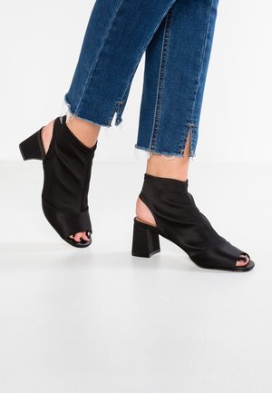 Fødder iført sorte sko med åbne tæer og åbne hæle, blokhæl, ankelhøj strækstof, kombineret med blå jeans med frynset kant og små slidser.