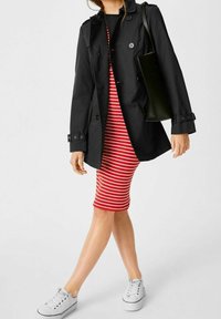 Trench-coat noir à boutons avec taille ceinturée, porté sur une robe ajustée rayée rouge et blanche, associé à des baskets blanches.