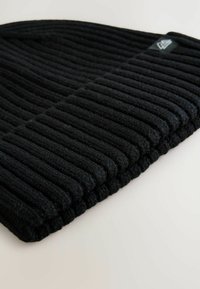 Zwarte geribbelde breiver van een textuurstof. Voorzien van een omgeslagen rand en een klein merklabel aan de zijkant. Zacht en dikke materiaal.