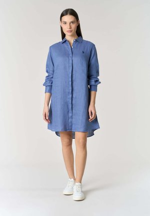 Donna dai capelli lunghi e scuri che indossa un vestito camicia blu con bottoni e sneakers bianche, in piedi contro uno sfondo chiaro e uniforme.