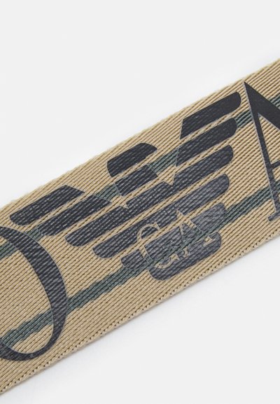 Sangle en tissu beige avec un logo d'aigle noir et des rayures horizontales vertes. La texture est côtelée avec un léger éclat.
