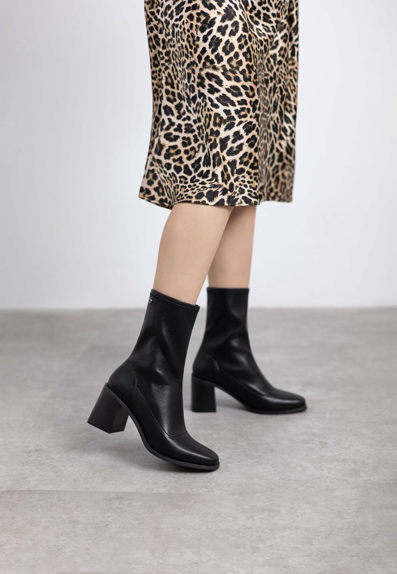 Bottines en cuir noir avec des orteils carrés et des talons épais, associées à une jupe en imprimé léopard. Texture lisse et design ajusté.