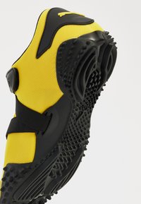 Yellow mesh athletic shoe with black accents, Velcro strap, and textured rubber sole featuring nubs for traction and grip.
