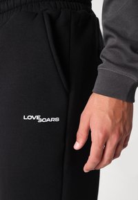 Melnas sporta bikses ar aukliņu jostiņu, ar baltu "LOVE SCARS" logo uz augšstilba. Mīksta tekstūra, eleganta forma un sānu kabatas.