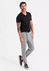 T-shirt noir à col en V, pantalons plissés gris clair, chaussures décontractées grises, bras tatoué visible, debout devant un fond blanc.