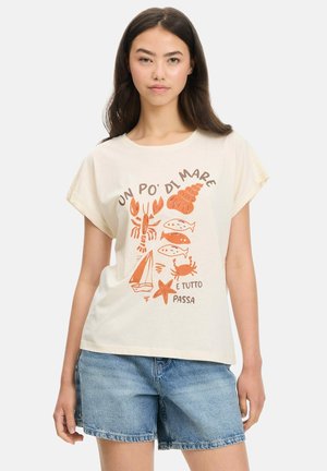 Mujer que lleva una camiseta blanca con ilustraciones naranjas con temática marina y texto en italiano, combinada con pantalones cortos vaqueros azules, de pie sobre un fondo blanco.