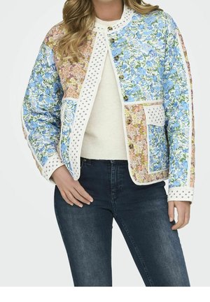 Vrouw draagt een patchwork jas met bloemenmotief in blauw en roze, met een bies met stippen, bruine knopen, een crèmekleurige trui en donkerblauwe spijkerbroek.