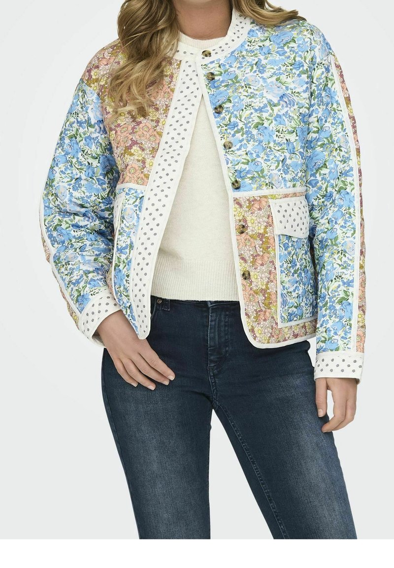 Femme portant une veste patchwork fleurie avec des fleurs bleues et roses, bordure à pois, boutons marron, pull crème et jean bleu foncé.