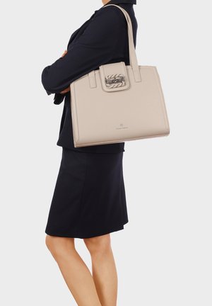 Femme en tailleur noir portant un grand sac à main en cuir beige avec un détail de boucle argentée sur l'épaule.