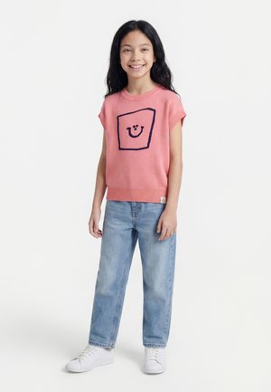 Jeune fille aux longs cheveux noirs portant un pull sans manches rose avec un motif de visage souriant, un jean bleu clair et des baskets blanches, debout.