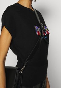 Schwarzer, kurzärmliger, gerippter Pullover mit farbenfrohem Perlen-Logo-Detail. Kombiniert mit einer schwarzen Schultertasche mit silbernen Beschlägen.