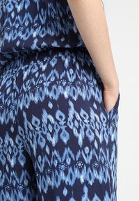 Granatowy materiał z niebieskim wzorem ikat i abstrakcyjnymi kształtami. Elastyczny pas z bocznymi kieszeniami i miękką, lekką teksturą.