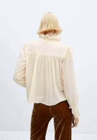 Blusa color crema con colletto a volant, maniche lunghe e tessuto strutturato. Presenta un dettaglio arricciato sul retro e polsini con bottoni.