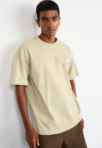 Beige cotton t-shirt with a relaxed fit, featuring short sleeves and a small cloud graphic embroidered on the left chest.