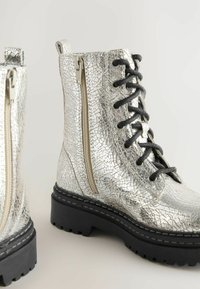 Bottines métalliques argentées avec une texture craquelée, lacets noirs et zip sur le côté. Semelle en caoutchouc noir épais avec traction.