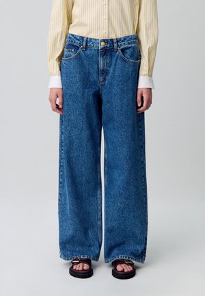 Jean en denim à jambes larges dans un délavage bleu moyen. Comprend deux poches avant, une fermeture à bouton en métal et une taille haute. Texture lisse et ourlets cousus.