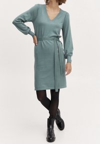 Abito a maglia a V in teal con maniche lunghe, vita con cintura e orlo a ginocchio, abbinato a stivaletti neri alla caviglia. Texture liscia e design aderente.