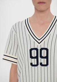 Personne portant un pull blanc à col en V manches courtes avec de fines rayures verticales noires et le numéro 99 en noir gras à l’avant.