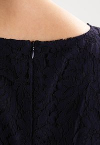 Décolleté d'une robe en dentelle noire avec des motifs floraux délicats. Fermeture à glissière dissimulée au dos. Tissu texturé avec des superpositions transparentes.