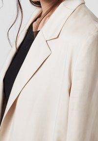 Světle béžový blazer z hladkého materiálu, s klopovým límcem, dlouhými rukávy a přiléhavým střihem, nošený přes černý top.