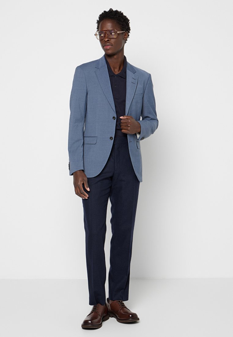 Mann in blauer Blazerjacke, dunkelblauer Hemd, dunkelblauen Hosen, braunen Oxfords und klaren Brillengestell, stehend vor einem weißen Hintergrund.