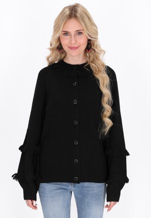 Blonde woman wearing black buttoned sweater with fringe details on sleeves and collar, paired with light blue jeans, smiling at camera.