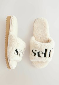Fuzzy cremede slippers med sort tekst, blød overdels, åben tå-design og kontrasterende tekstureret sål. Blødt materiale og afrundet form.