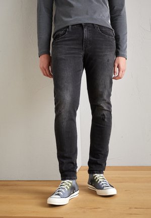 Jeans de denim negro con corte ajustado, que presentan un lavado deslavado, desgaste en las rodillas y bolsillos traseros cosidos, combinados con zapatillas azules.