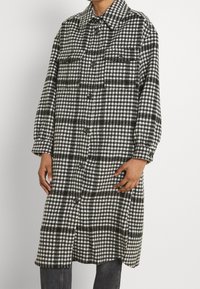 Přehledná černobílá oversized košile-jacket s knoflíky vpředu, dvěma náprsními kapsami a límcem. Vyrobena z měkké tkaniny.