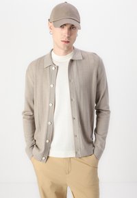 Μπεζ κάρντιγκαν με επίπεδο γιακά, κλείσιμο με κουμπί και υφή κατακόρυχες ρίγες, τοποθετημένο πάνω από άσπρη μπλούζα με στρογγυλή λαιμόκοψη και μπεζ παντελόνι.