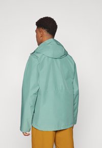 Chaqueta resistente al agua en color verde azulado con capucha, de corte recto, mangas largas y textura suave. Llevada sobre pantalones de color marrón claro.
