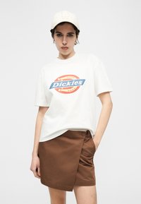 Νεαρή γυναίκα που φοράει λευκή μπλούζα με το λογότυπο "Dickies", καφέ φούστα τύπου wrap και λευκό καπέλο μπέιζμπολ με το λογότυπο LA, ποζάροντας με το ένα χέρι στην τσέπη της φούστας.