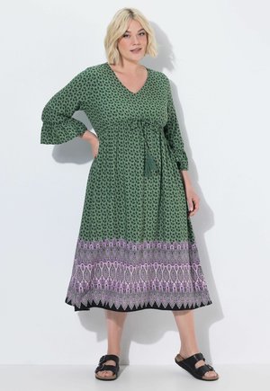 Lyshåret kvinde iført en grøn midi-kjole med små sorte blomsterprint og lilla mønstret kant, kombineret med sorte sandaler med spænde.