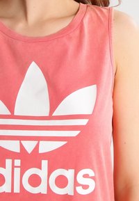 Rosa ärmlös tanktopp med stort vitt Adidas-logotyp med trebladig design och horisontella ränder under varumärket. Mjuk bomullsmaterial.