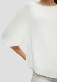 Blouse blanche plissée avec un large col bateau, en tissu à texture douce, et des manches longues bouffantes et amples. Conçue avec une coupe décontractée.