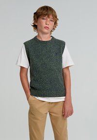 Vihreä ilmanhiha knit-neule, jossa on teksturoitu kuvio, päällä valkoinen t-paita, yhdistettynä beigeihin chinokapan. Neuleessa on ribbireuna.