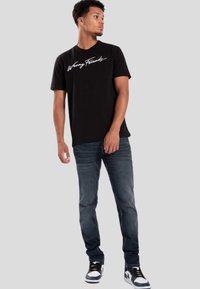Camiseta negra de algodón con el texto blanco "Amigos Equivocados", combinada con jeans azul oscuro y zapatillas negras y blancas. El modelo está de pie, mirando hacia la izquierda.