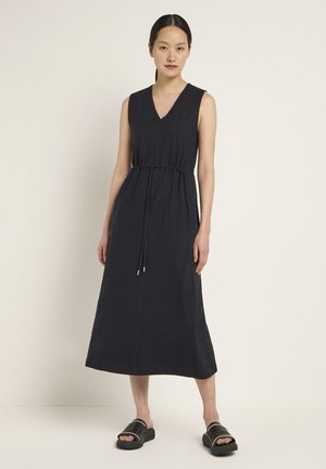 Ärmelloses Maxikleid mit V-Ausschnitt aus dunkelgrauer Baumwolle. Verfügt über eine verstellbare Kordelzugtaille und eine gerade Silhouette. Flauschige Sandalen getragen.