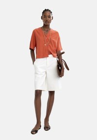 Pomarańczowa koszula z krótkim rękawem zapinana na guziki, białe szorty do kolan, czarne płaskie buty i brązowa torebka. Prosty detail z warstwowym naszyjnikiem.