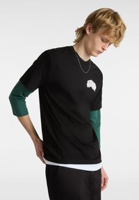 Camiseta de algodón negra de manga corta sobre una camisa de manga larga verde. Presenta un pequeño gráfico blanco en el pecho y un look en capas.