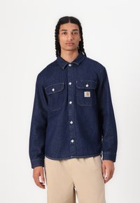 Dark denim shirt with two chest pockets, snap buttons, and a small logo patch. Features a collar and long sleeves, with contrast stitching.