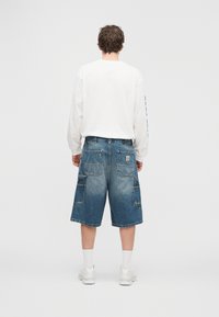 Τζιν σορτς με μεσαία πλύση, πολλαπλές τσέπες και ταμπέλα με το λογότυπο Carhartt. Συνδυασμένα με λευκό πουκάμισο με μακριά μανίκια και αθλητικά παπούτσια.